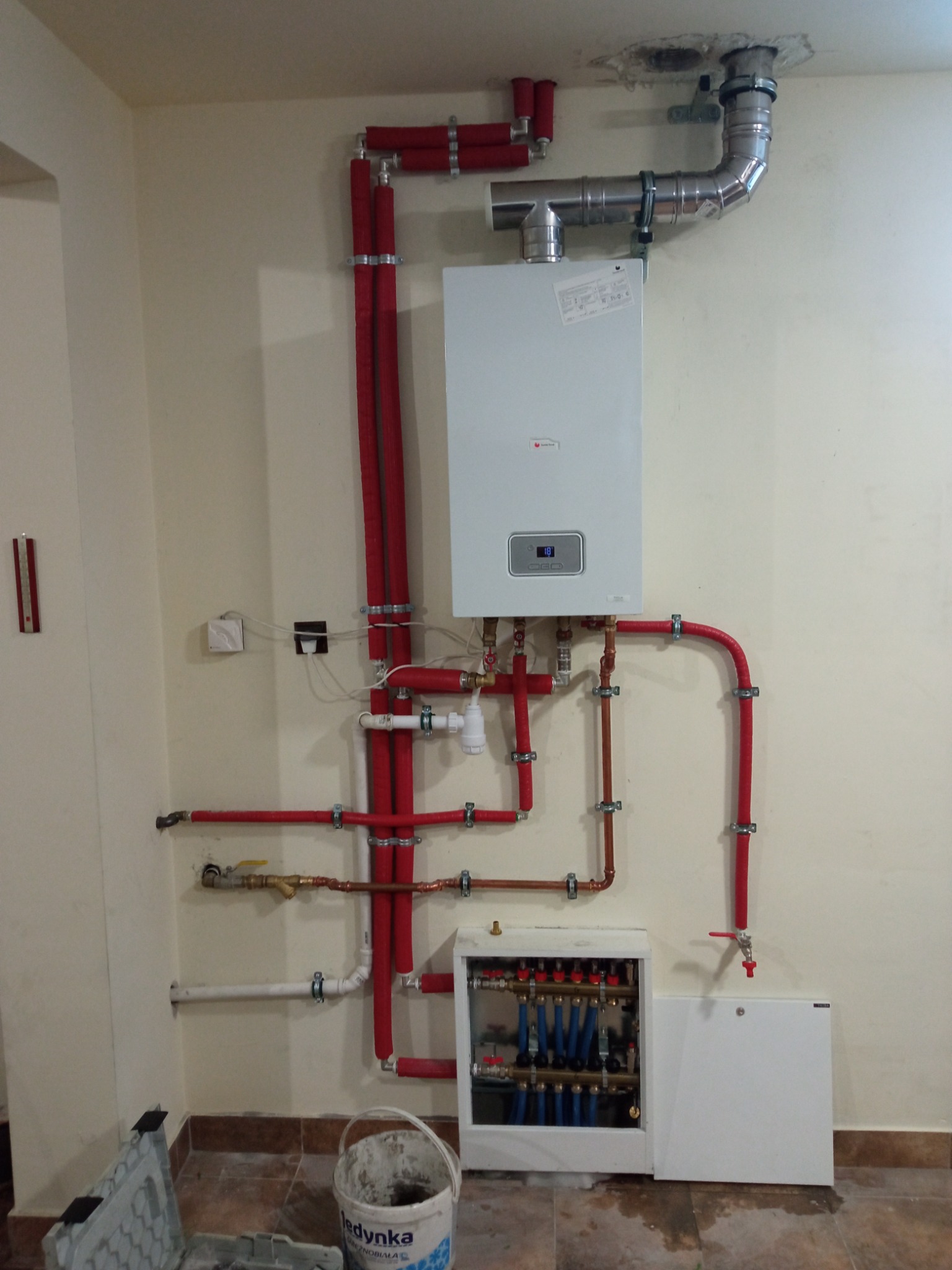 Biały piec gazowy zamontowany na ścianie z podłączonymi rurami grzewczymi w izolacji czerwonej, rozdzielacz ogrzewania podłogowego z niebieskimi pętlami widoczny poniżej, ślady po zalaniu na podłodze.