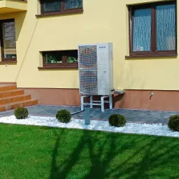 Biała, dwuczęściowa zewnętrzna jednostka klimatyzacji Samsung na metalowym stelażu, umieszczona na szarym chodniku z białym żwirem, przed żółtym budynkiem z brązowymi oknami i zielonym trawnikiem...