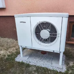 Zewnętrzna jednostka pompy ciepła Stiebel Eltron na betonowych płytach z białym żwirem, na tle ściany budynku z małym oknem.