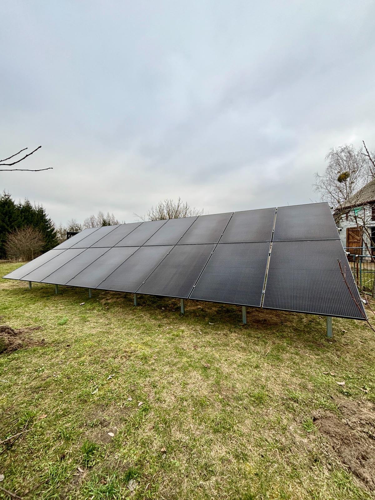 Panel fotowoltaiczny na metalowej konstrukcji, ustawiony na trawniku przed domem z drewnianymi okiennicami, pochmurne niebo w tle.
