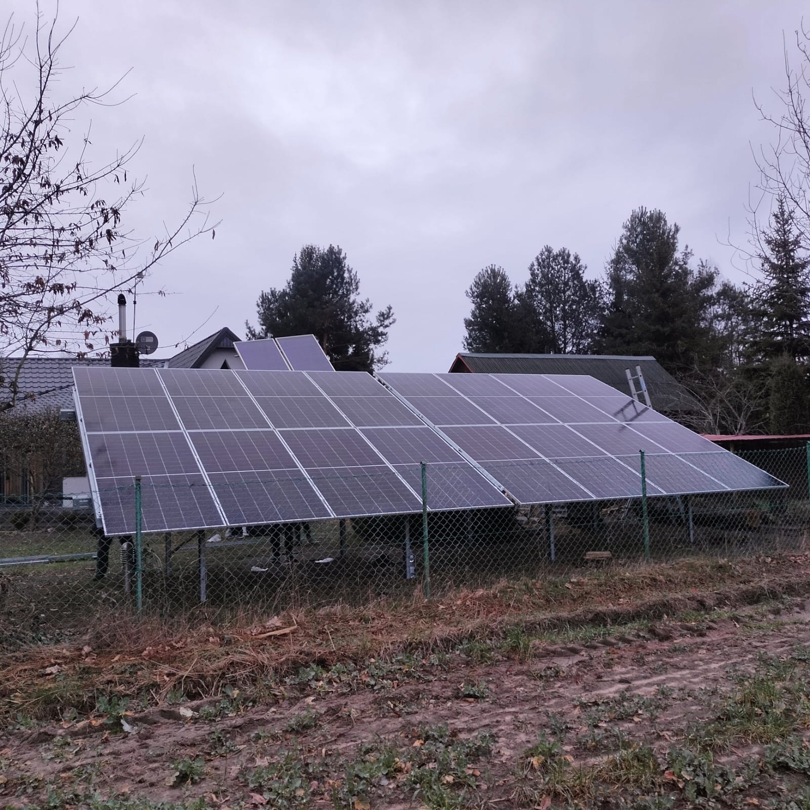 Panel fotowoltaiczny zamontowany na gruncie za ogrodzeniem z siatki, widok na dom w tle, pochmurny dzień.