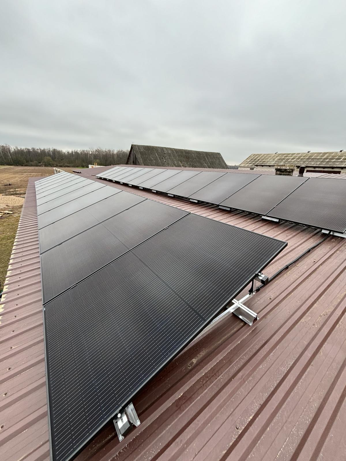 Panele słoneczne zamontowane na metalowym dachu budynku gospodarczego, widoczne elementy montażowe i okablowanie, pochmurne niebo w tle.