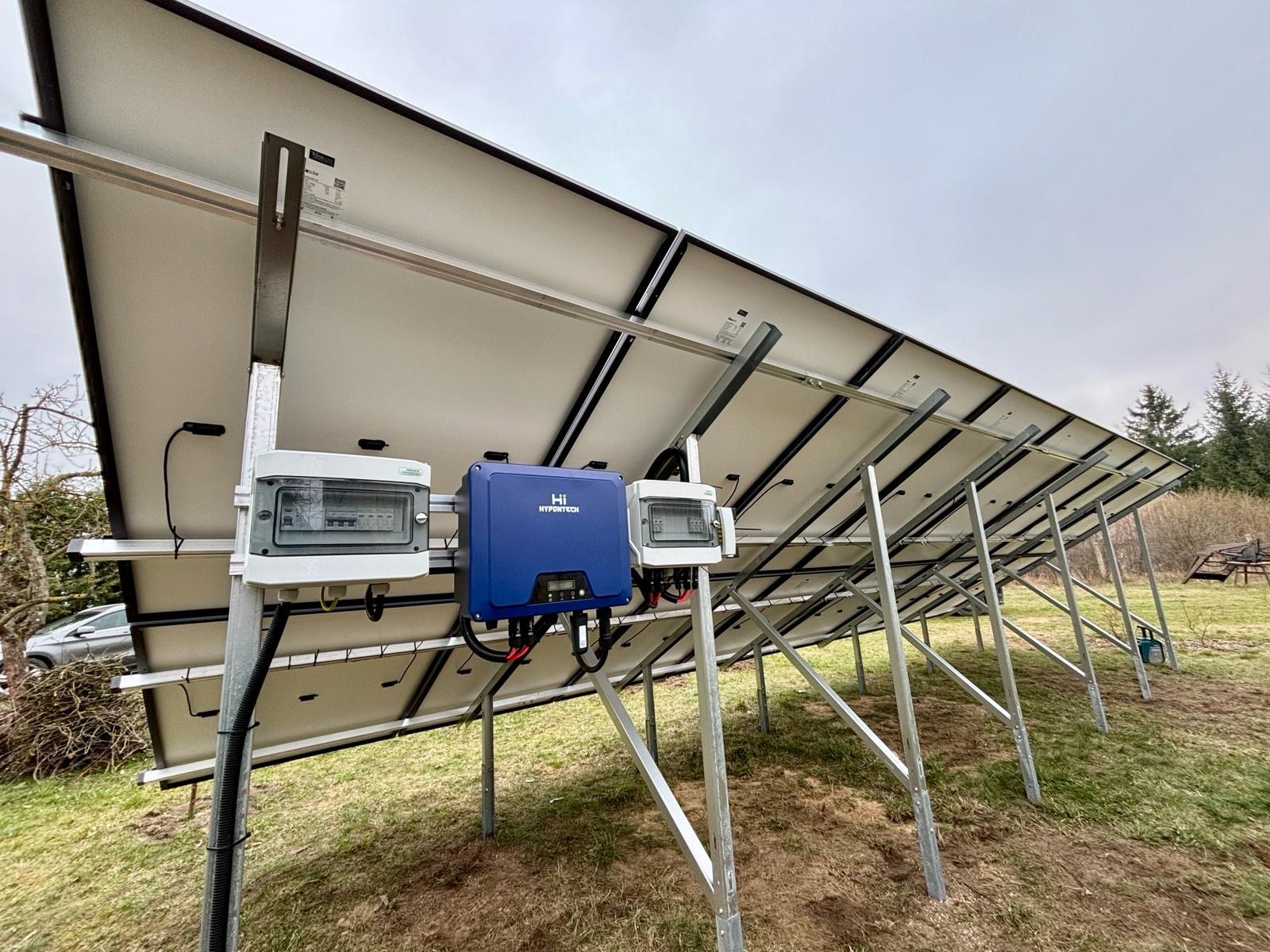 Montaż paneli fotowoltaicznych widziany od spodu, z widocznymi skrzynkami elektrycznymi i inwerterem Hypotech na tle pochmurnego nieba i trawy.