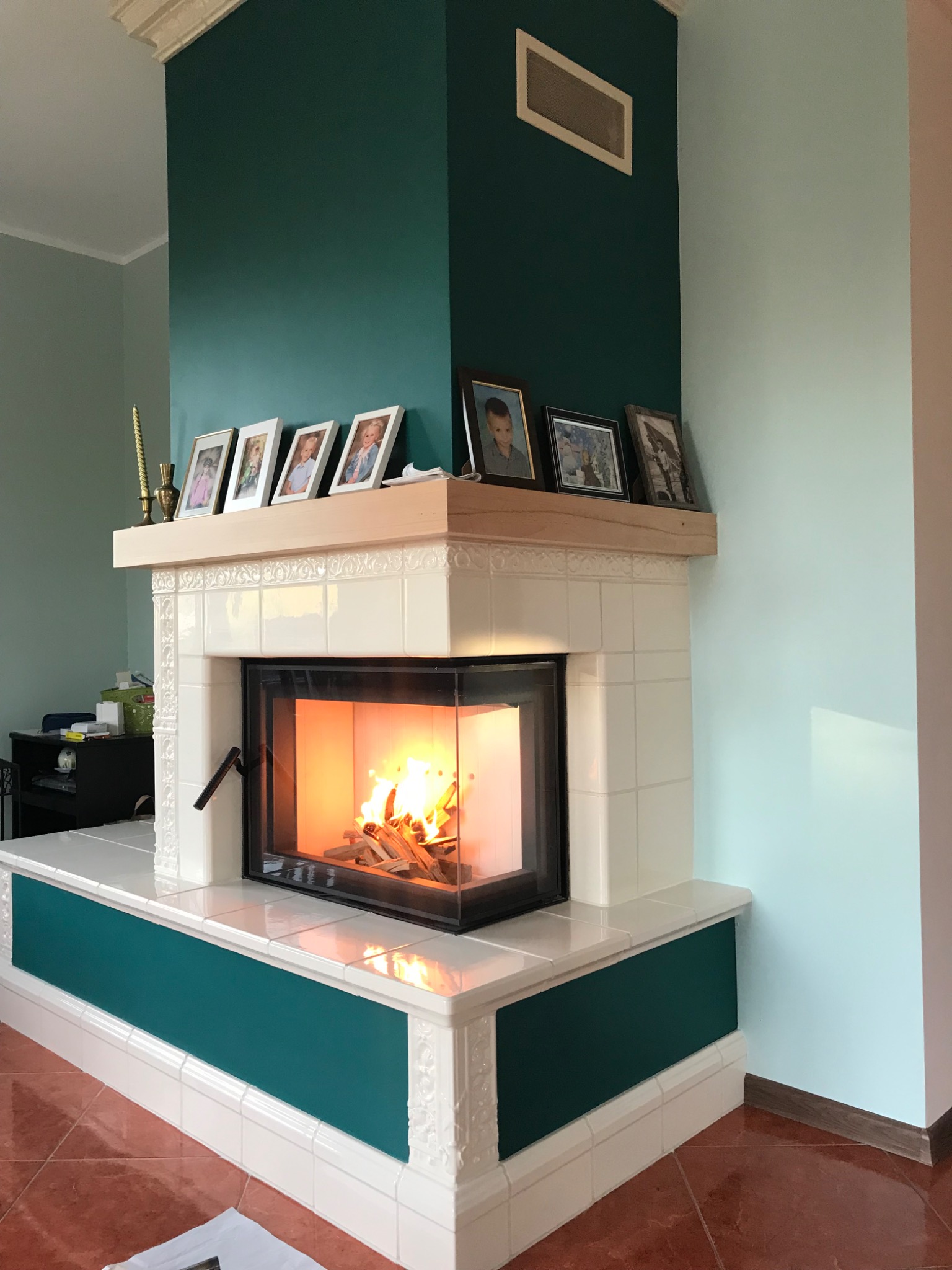Biały kominek z paleniskiem i czarną ramą, obudowany płytkami, z drewnianym blatem, na którym stoją ramki ze zdjęciami. Ściany w odcieniach zieleni.