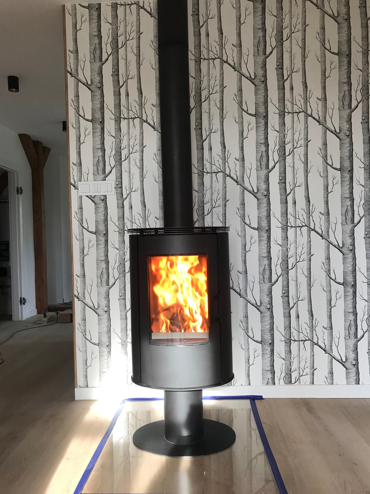 Nowoczesny, czarny piec wolnostojący z płomieniami, na tle ściany z motywem drzew. Podłoga zabezpieczona folią i taśmą malarską. Ujęcie centralne.