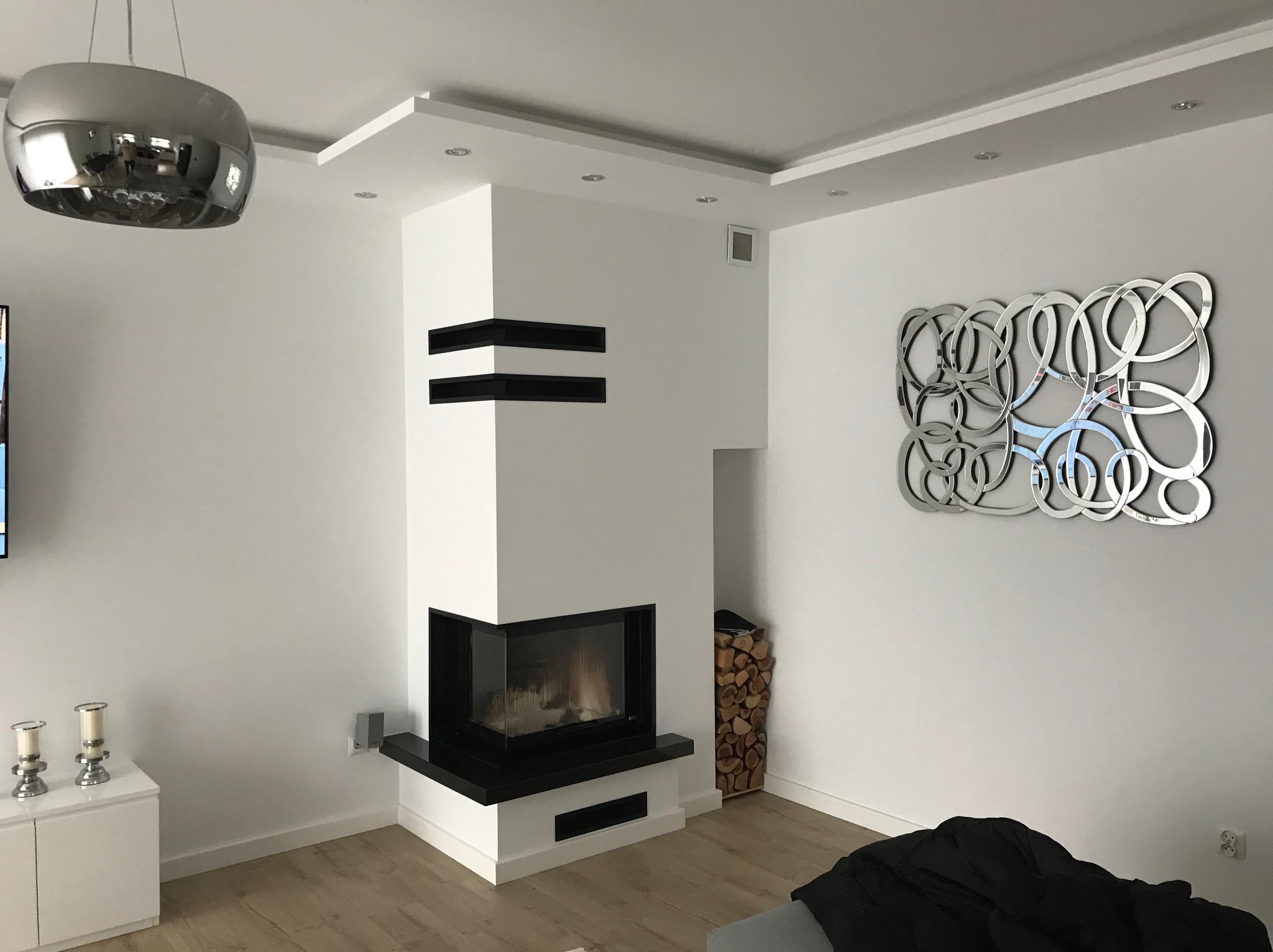 Nowoczesny, narożny wkład kominkowy z czarnymi akcentami w jasnym salonie. Drewno ułożone obok, srebrna dekoracja na ścianie, designerska lampa sufitowa.
