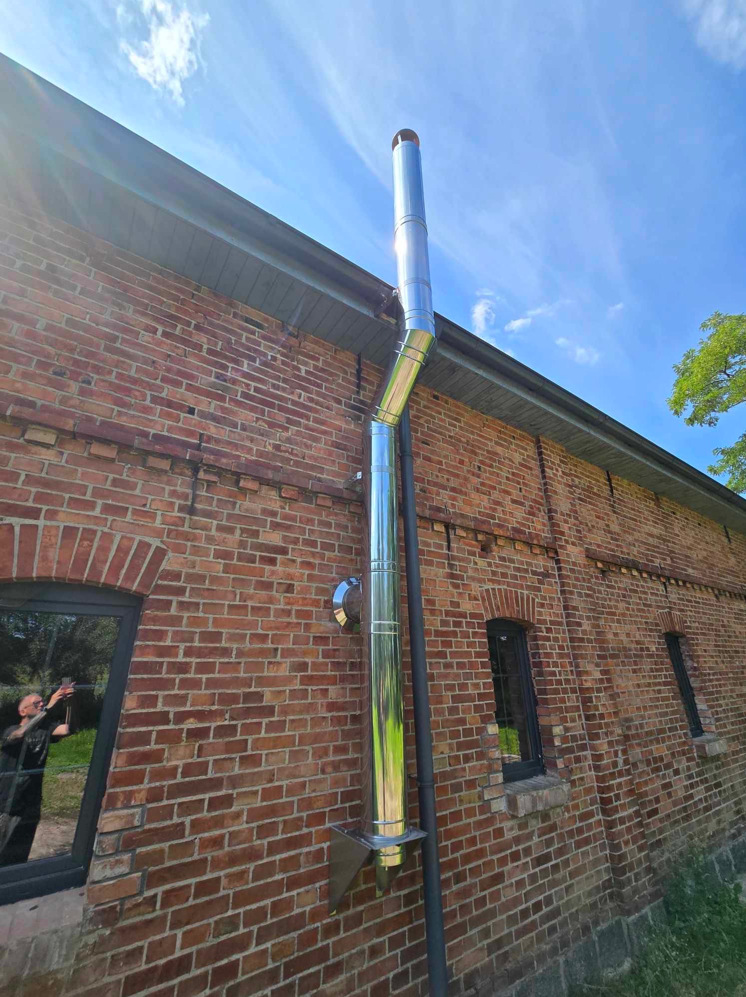 Zewnętrzny, stalowy przewód kominowy zamontowany na ceglanej ścianie budynku. Widoczne elementy mocujące i odbicie osoby w oknie. Błękitne niebo w tle.