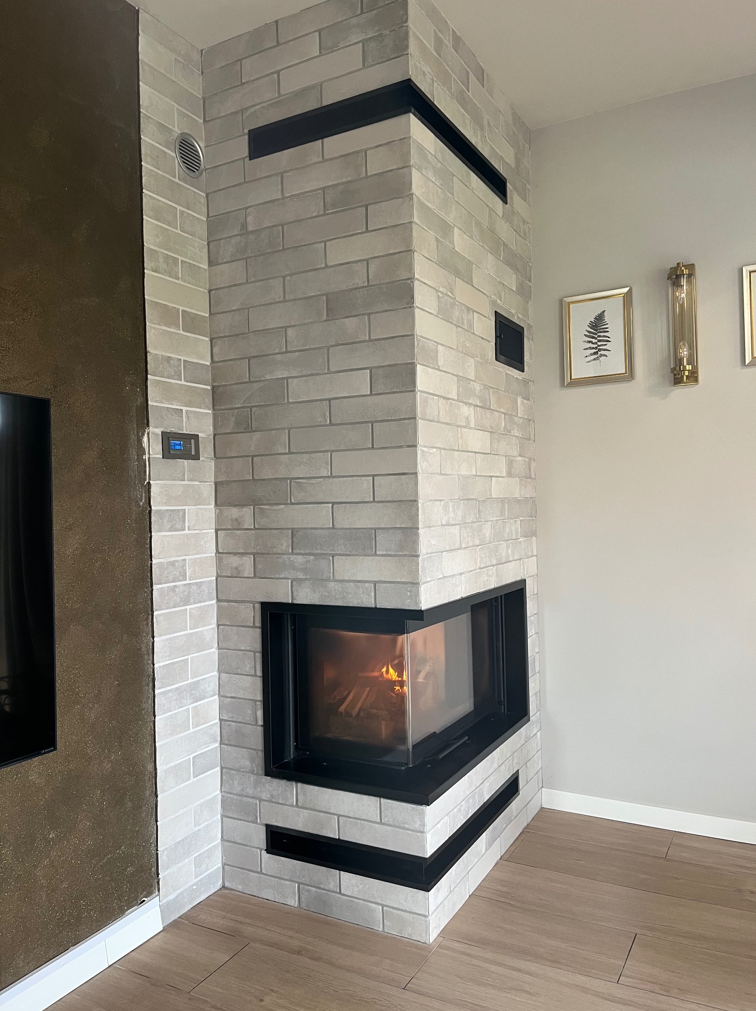 Narożny, nowoczesny  z obudową z białej cegły i czarnymi akcentami. W palenisku widoczny ogień. Na ścianie dekoracje i oświetlenie. Drewnopodobna podłoga.