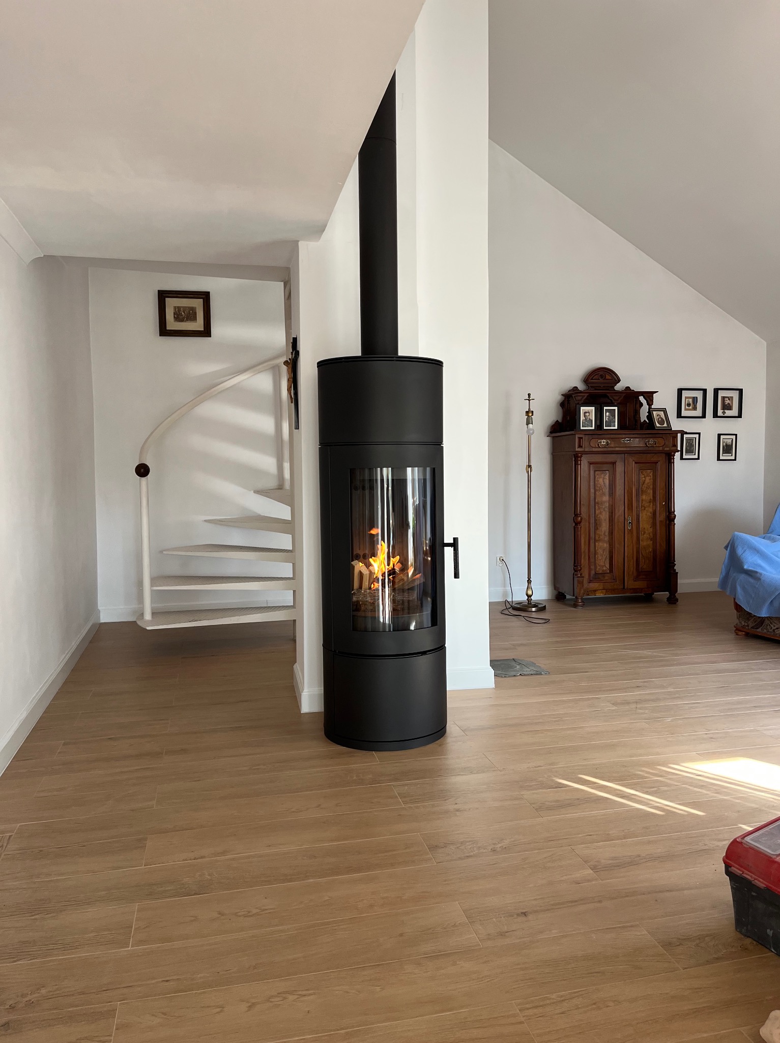Nowoczesny, czarny piec wolnostojący z płomieniami, oddzielający spiralne schody od salonu z drewnianą podłogą i antycznym kredensem. Jasne wnętrze.