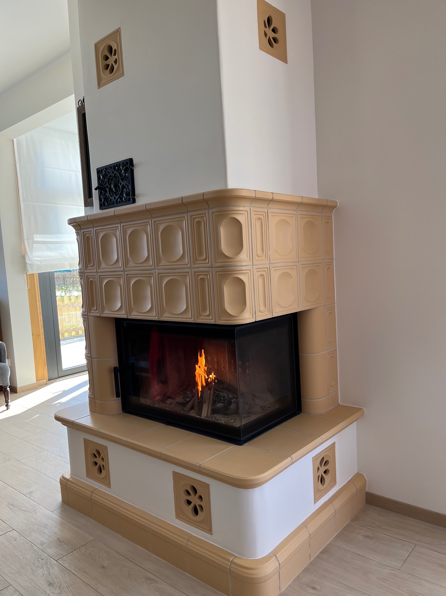 Nowoczesny kominek kaflowy z paleniskiem, wykończony beżowymi płytkami z motywem kwiatowym, w jasnym wnętrzu. Widoczne płomienie i dekoracyjne kratki wentylacyjne.