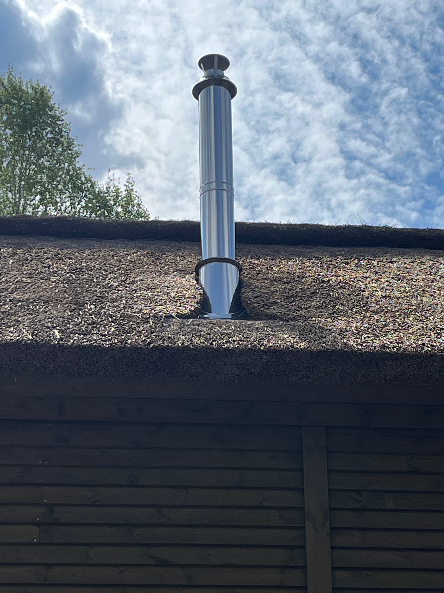 Błyszczący, metalowy przewód kominowy na strzechą pokrytym dachu, widziany z dołu na tle błękitnego nieba z chmurami. Ciemne, drewniane panele w dolnej części kadru.