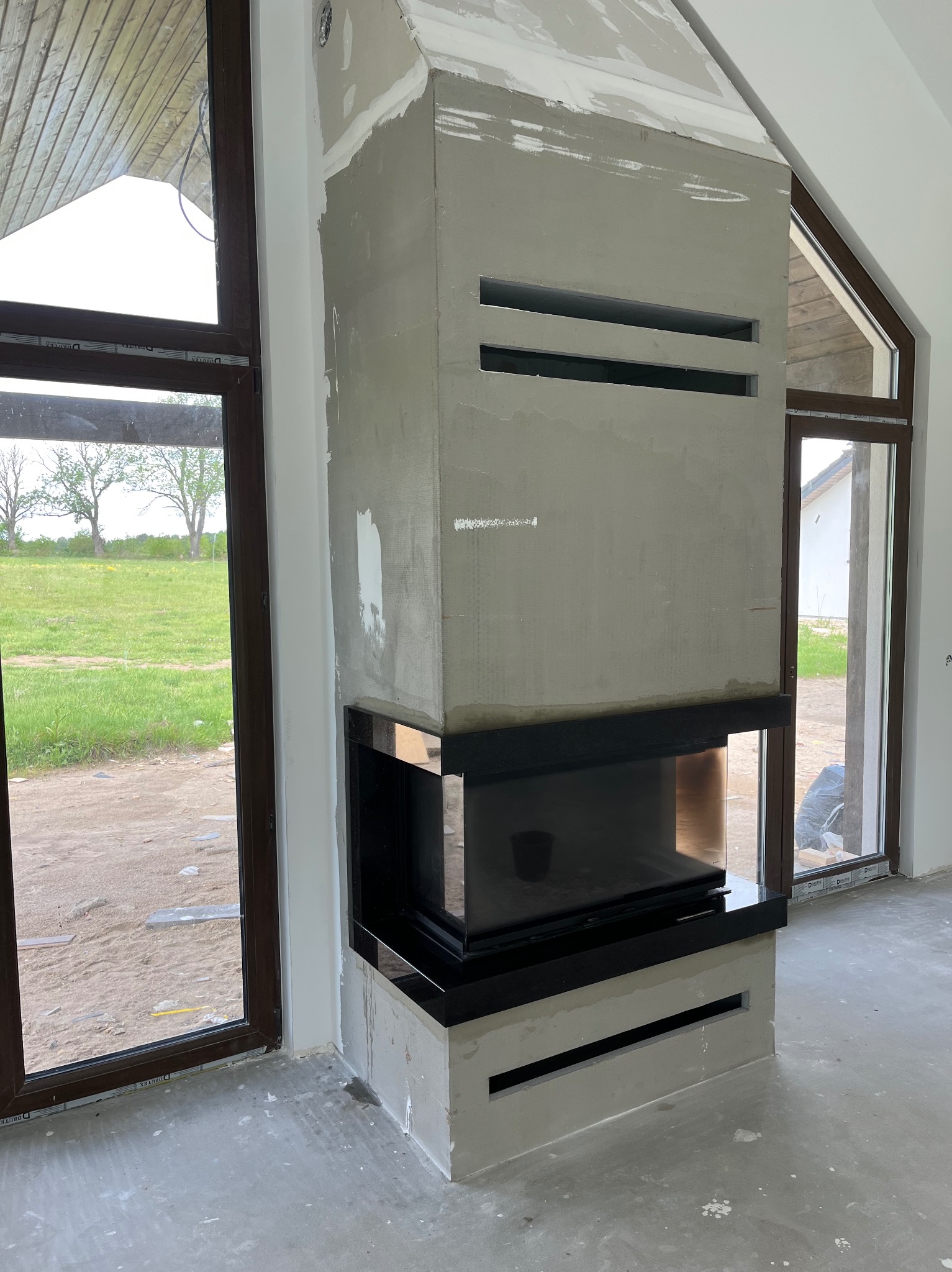 Nowoczesny, narożny wkład kominkowy z czarnym obramowaniem, w trakcie obudowy z szarego betonu architektonicznego. Widoczne okna i surowa podłoga.