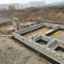 Wznoszenie ścian fundamentowych z bloczków betonowych na mokrym placu budowy, widoczne pionowe pręty zbrojeniowe i niebieskie rury instalacyjne.