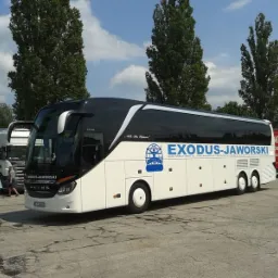 Biało-czarny autokar firmy Exodus-Jaworski zaparkowany na betonowym placu w słoneczny dzień, z widocznym logiem firmy i tablicą rejestracyjną. W tle drzewa i zaparkowane naczepy TIR.