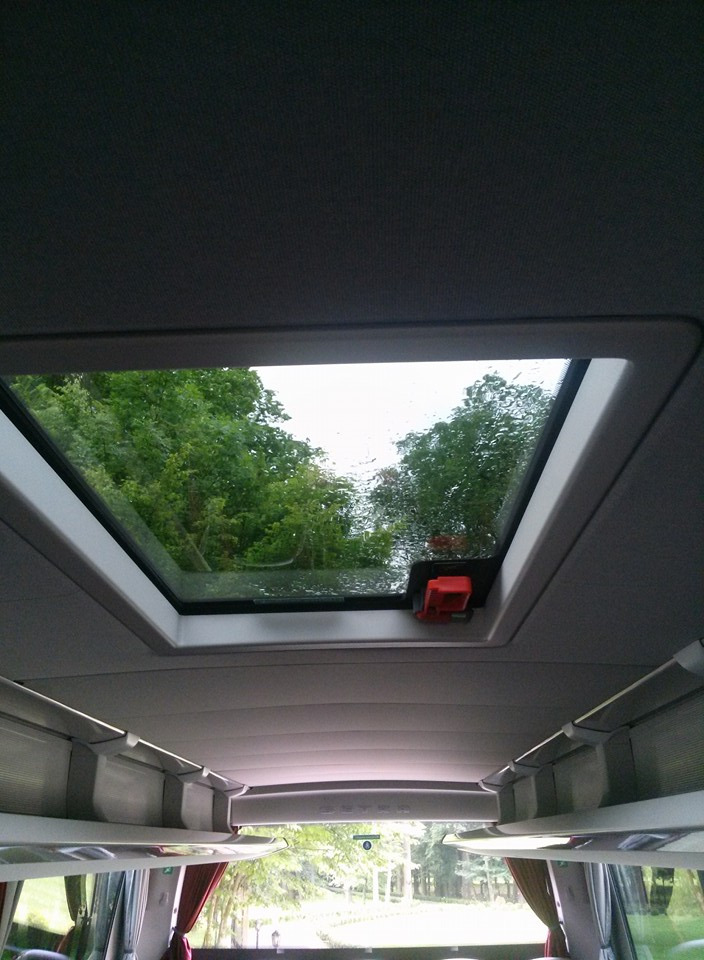 Wnętrze autobusu z widokiem przez otwierany świetlik dachowy w deszczowy dzień, z widocznymi drzewami na zewnątrz.
