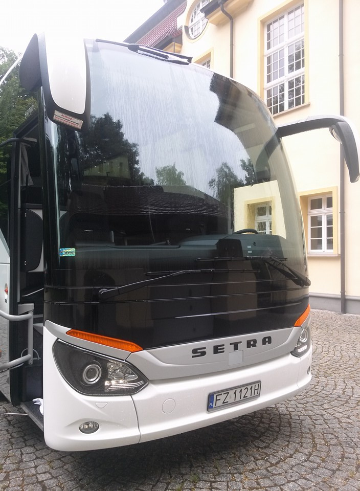 Przód nowoczesnego autokaru Setra z otwartymi drzwiami, zaparkowanego na brukowanym placu przed budynkiem z jasną elewacją i oknami z białymi ramami. Na szybie widoczne odbicie drzew i dachu.