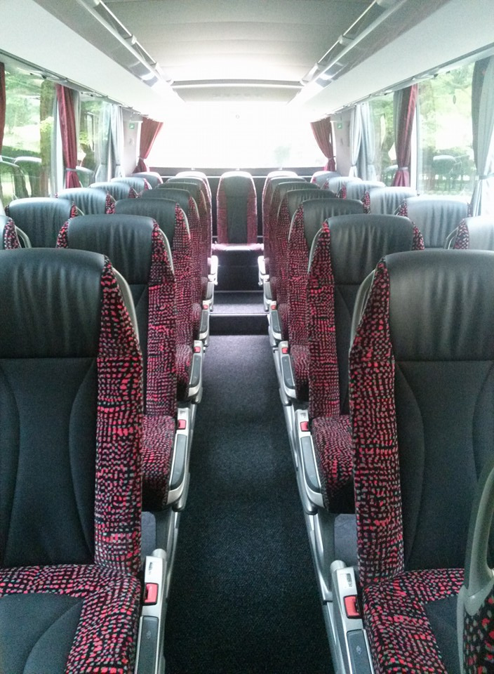 Wnętrze autobusu z rzędami czarnych foteli z czerwono-czarną tapicerką, widok z przodu pojazdu, ciemna podłoga, zasłony w oknach.