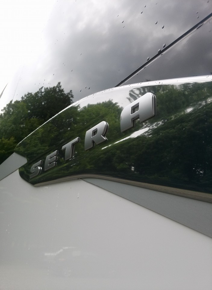 Błyszczący, chromowany napis SETRA na boku białego autobusu, z odbiciem zieleni drzew i kroplami deszczu na szybie.