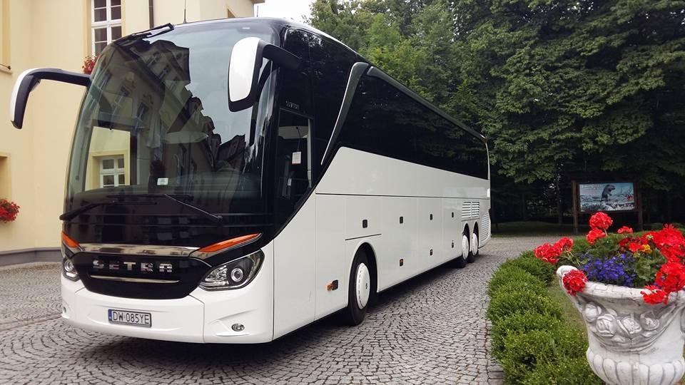 Nowoczesny biało-czarny autokar marki Setra zaparkowany na brukowanej drodze przed budynkiem z kwiatami w oknach i zielonymi drzewami w tle.