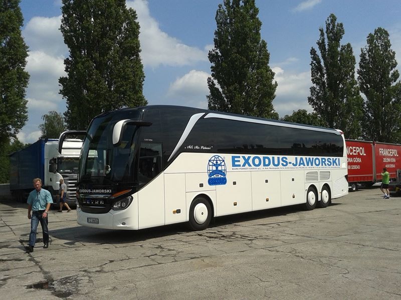 Biało-czarny autokar firmy Exodus-Jaworski zaparkowany na betonowym placu w słoneczny dzień, z widocznym logiem firmy i tablicą rejestracyjną. W tle drzewa i zaparkowane naczepy TIR.