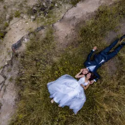 Para młoda leżąca na trawie w sukni ślubnej i garniturze, ujęcie z drona, widok z góry na tle skalistego terenu.