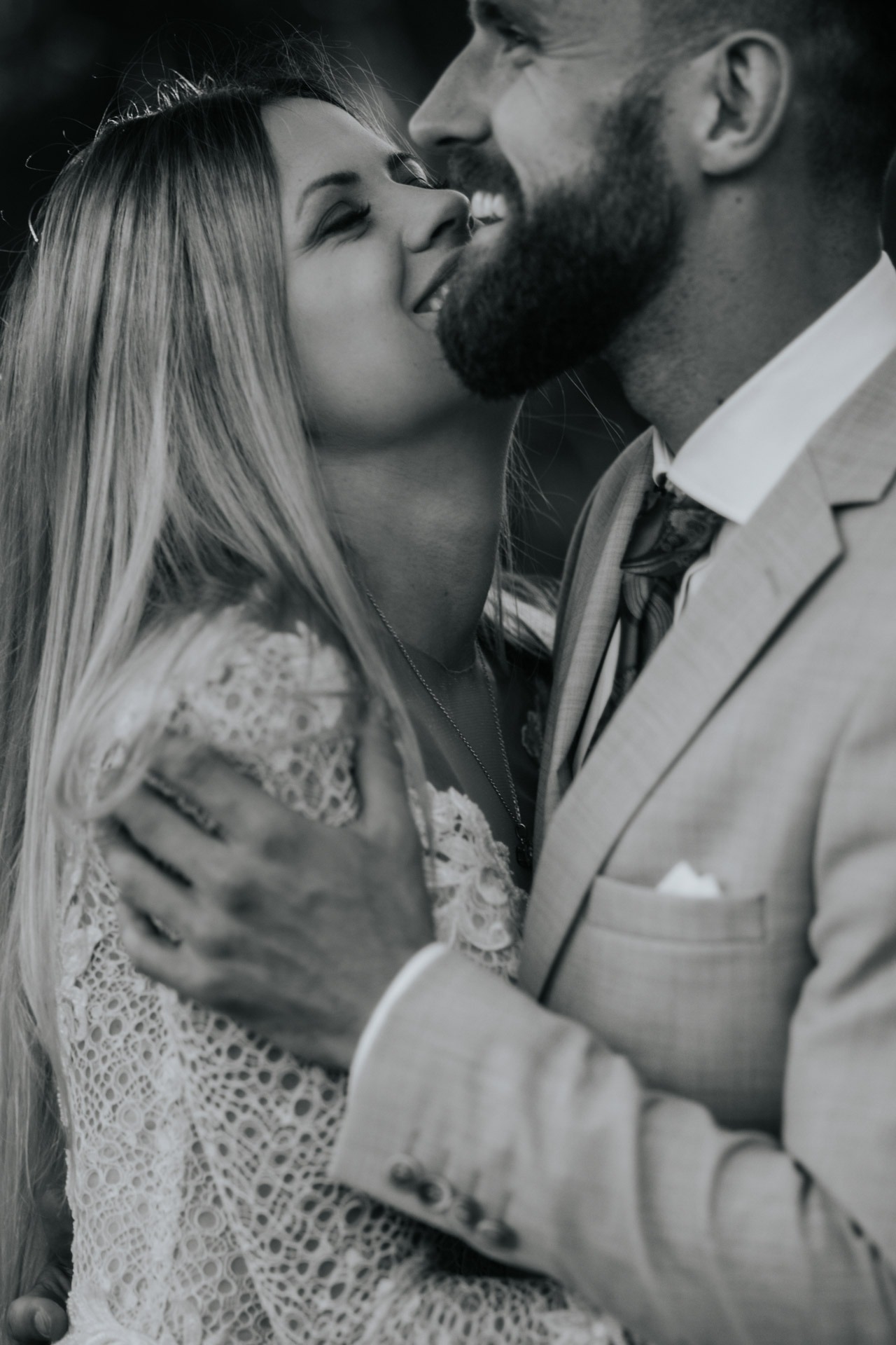 Czarno-białe zbliżenie uśmiechniętej pary: kobieta w koronkowej sukience z delikatnym naszyjnikiem i mężczyzna w garniturze z wzorzystym krawatem, obejmujący ją czule.