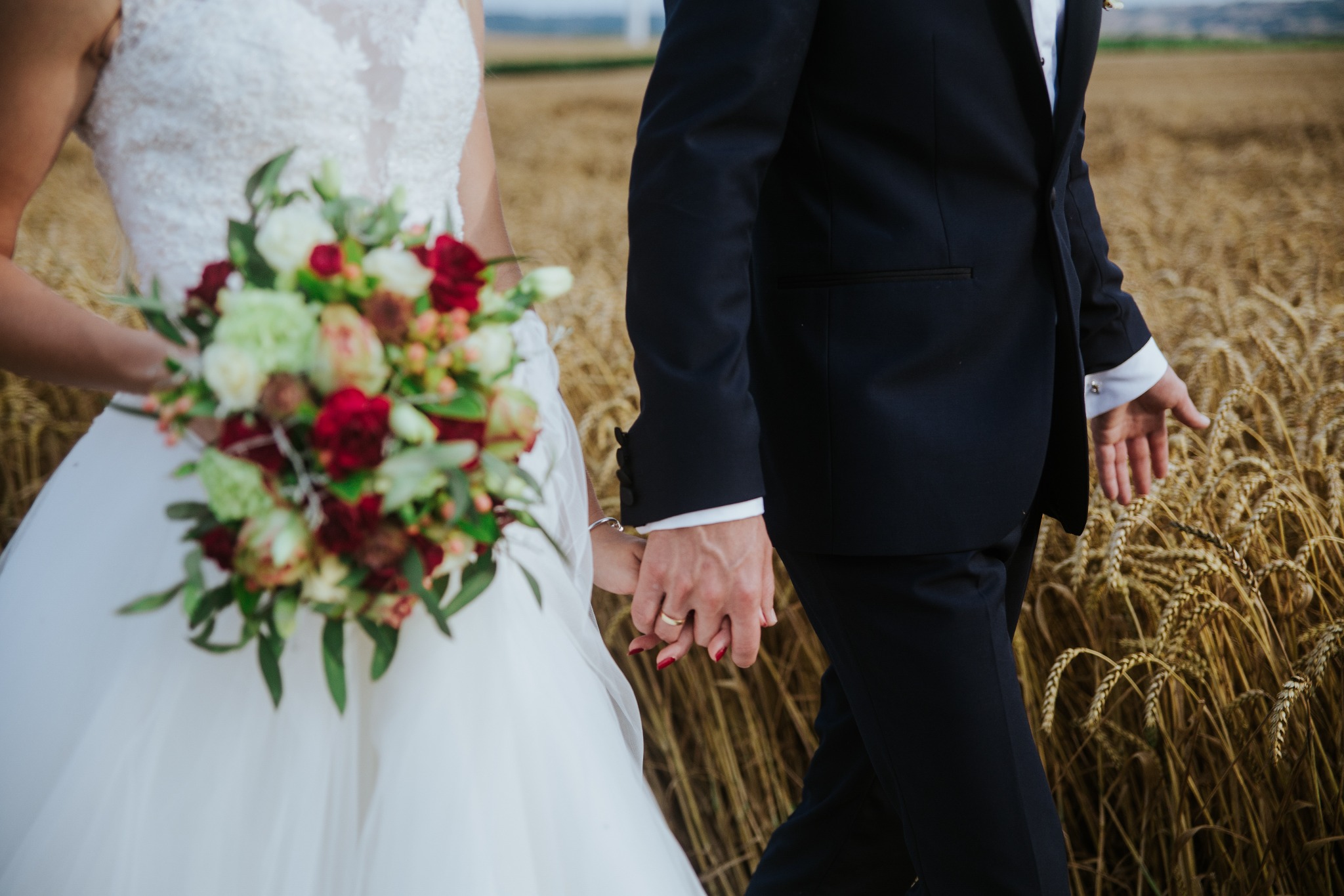 Para młoda trzymająca się za ręce na tle pola pszenicy. Panna młoda w białej sukni z bukietem czerwonych i białych kwiatów, pan młody w czarnym garniturze.