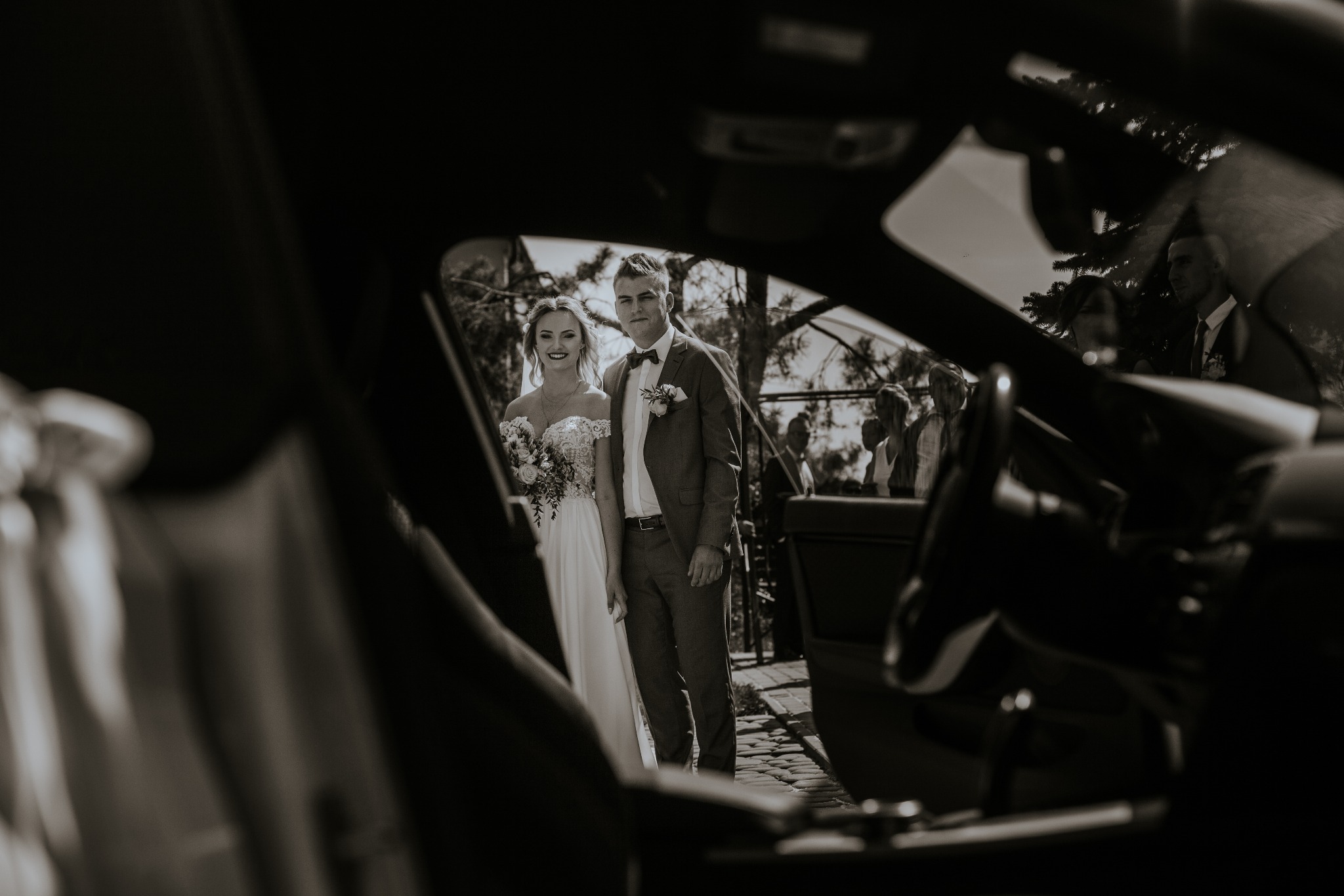 Para młoda w strojach ślubnych, widziana przez szybę samochodu z widocznym wnętrzem i kierownicą, na tle goście weselni i drzewa.