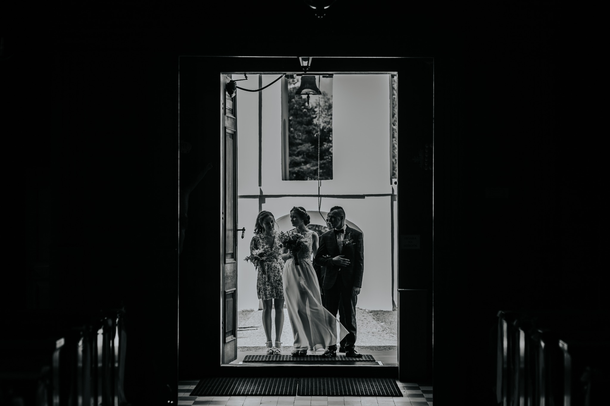 Czarno-białe zdjęcie panny młodej w koronkowej sukni, druhny z bukietem i mężczyzny w garniturze, stojących w drzwiach kościoła z dzwonem w tle.