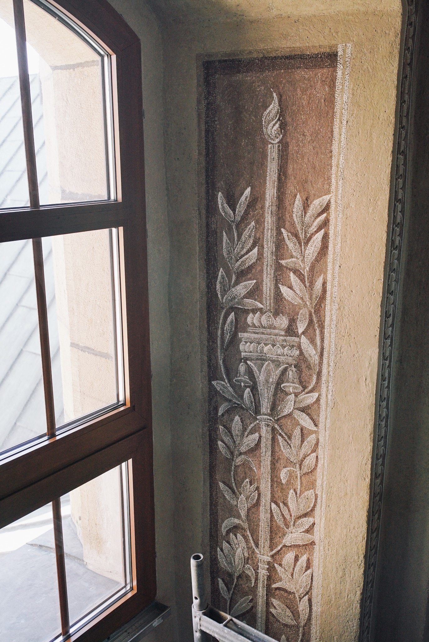 Fragment polichromii ściennej z motywem kandelabru i liści, widoczny obok drewnianego okna i fragmentu rusztowania.