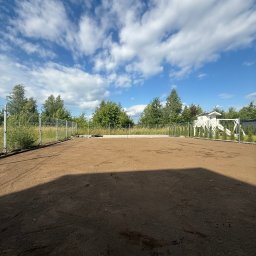 MM GROUP SPÓŁKA Z OGRANICZONĄ ODPOWIEDZIALNOŚCIĄ - Przygotowany teren pod trawnik z rolki, widoczne ślady opon i siatka ogrodzeniowa. W tle domek dla dzieci i błękitne niebo z chmurami.