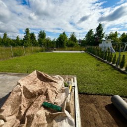 MM GROUP SPÓŁKA Z OGRANICZONĄ ODPOWIEDZIALNOŚCIĄ - Układanie trawy z rolki: widok na rozłożoną trawę, narzędzia pracy w przyczepce i przygotowany grunt. W tle drzewa i błękitne niebo z chmurami.