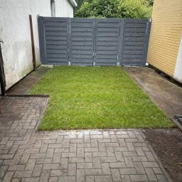 MM GROUP SPÓŁKA Z OGRANICZONĄ ODPOWIEDZIALNOŚCIĄ - Świeżo ułożona trawa z rolki w ogrodzie, ograniczona szarym płotem i kostką brukową. Widok z góry na fragment trawnika.