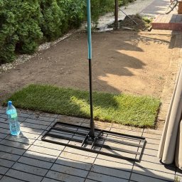 MM GROUP SPÓŁKA Z OGRANICZONĄ ODPOWIEDZIALNOŚCIĄ - Fragment układania trawy z rolki w ogrodzie, widoczny fragment rolki trawy, narzędzie do jej układania oraz butelka wody. W tle zielone tuje.