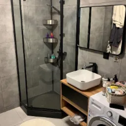 Nowoczesna łazienka z kabiną prysznicową w czarnej ramie, umywalką nablatową i pralką. Szare płytki i minimalistyczny design. Widoczny fragment lustra i akcesoria łazienkowe.