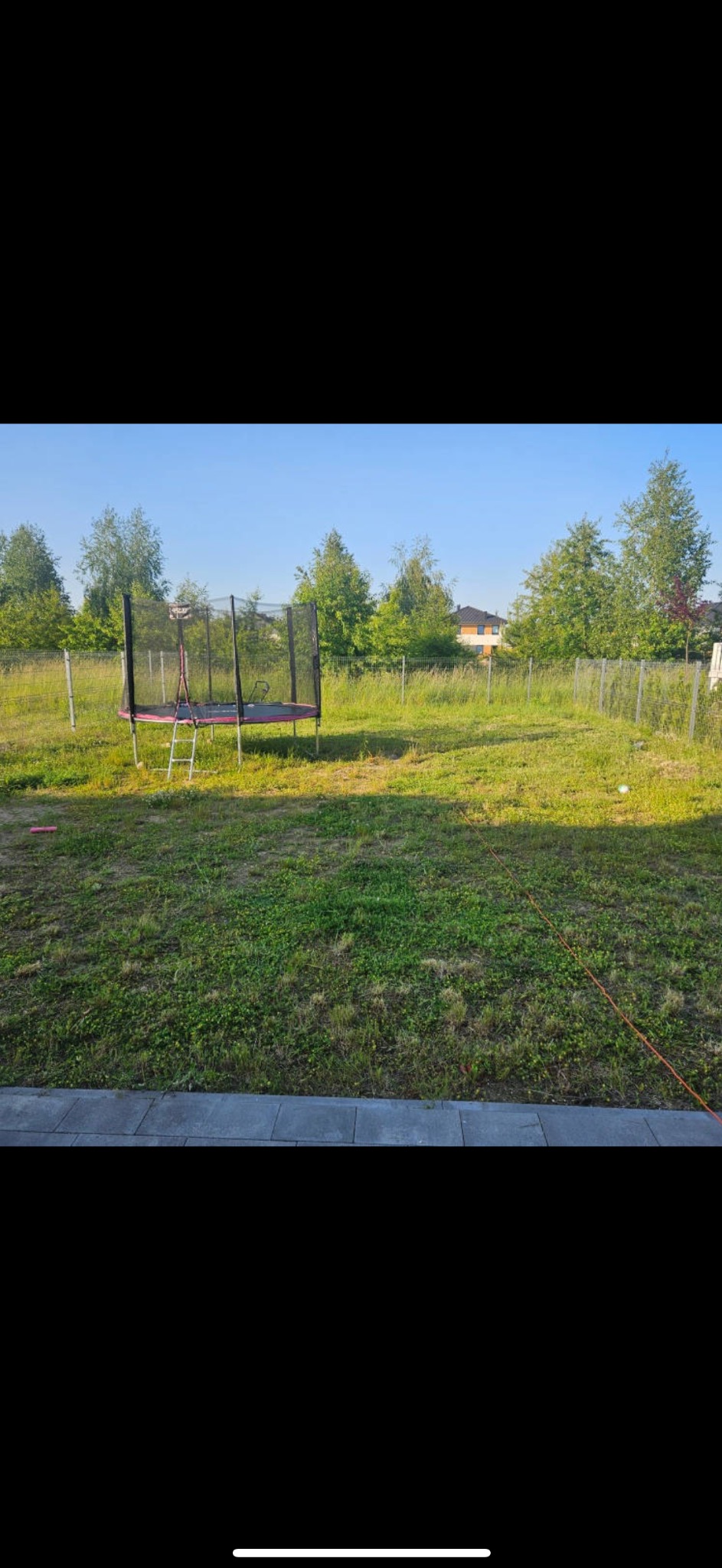 Zaniedbany trawnik z trampoliną, ogrodzony siatką. W tle drzewa i dom. Widok z tarasu z szarych płyt.