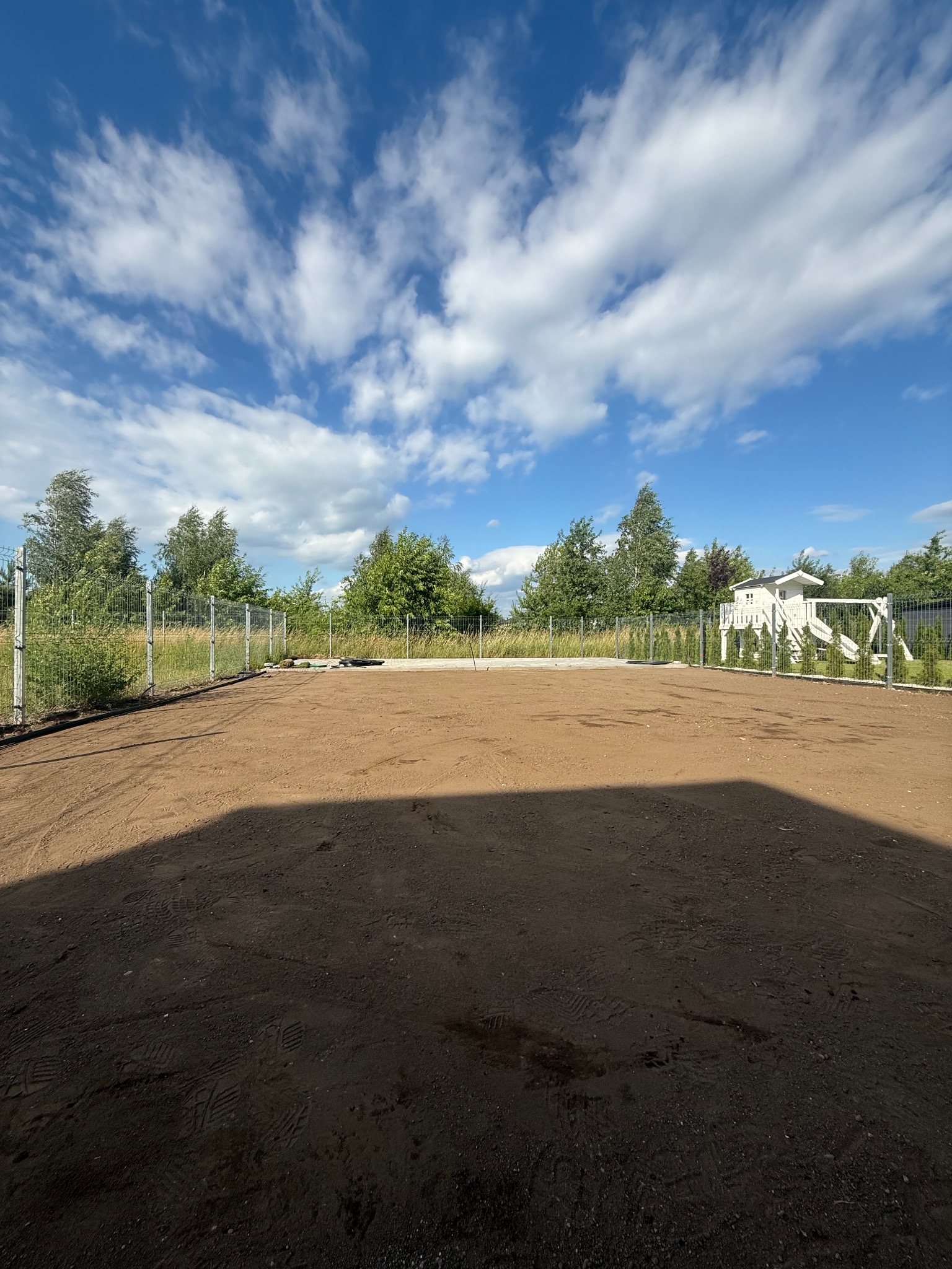 Przygotowany teren pod trawnik z rolki, widoczne ślady opon i siatka ogrodzeniowa. W tle domek dla dzieci i błękitne niebo z chmurami.
