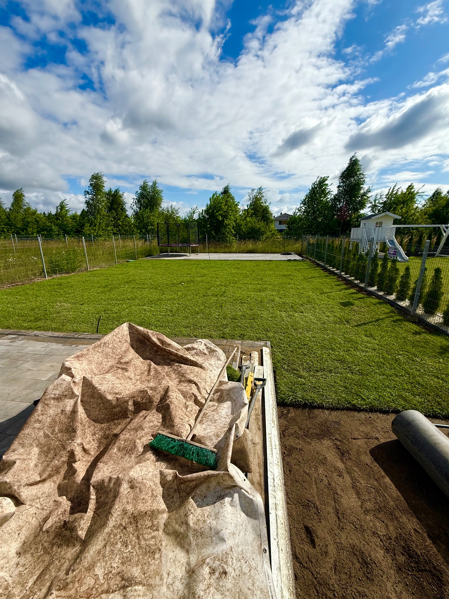 Układanie trawy z rolki: widok na rozłożoną trawę, narzędzia pracy w przyczepce i przygotowany grunt. W tle drzewa i błękitne niebo z chmurami.