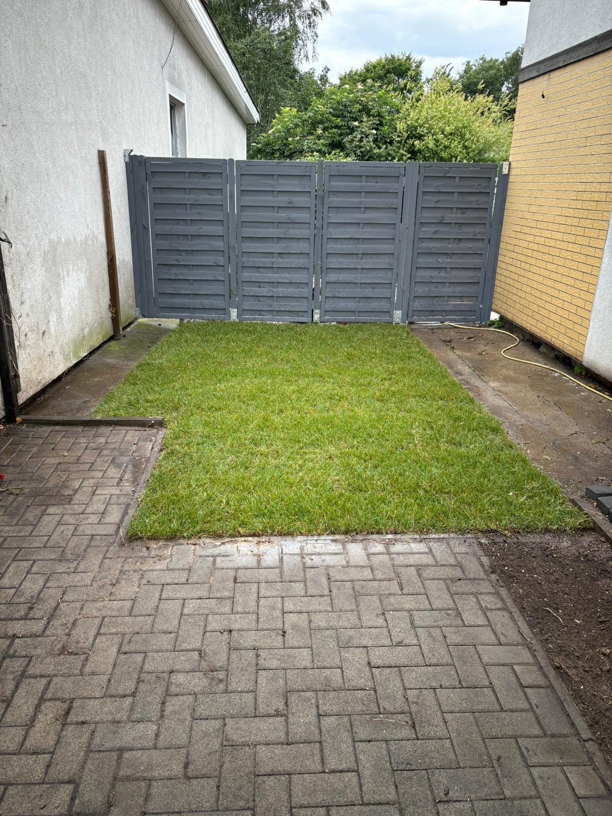 Świeżo ułożona trawa z rolki w ogrodzie, ograniczona szarym płotem i kostką brukową. Widok z góry na fragment trawnika.