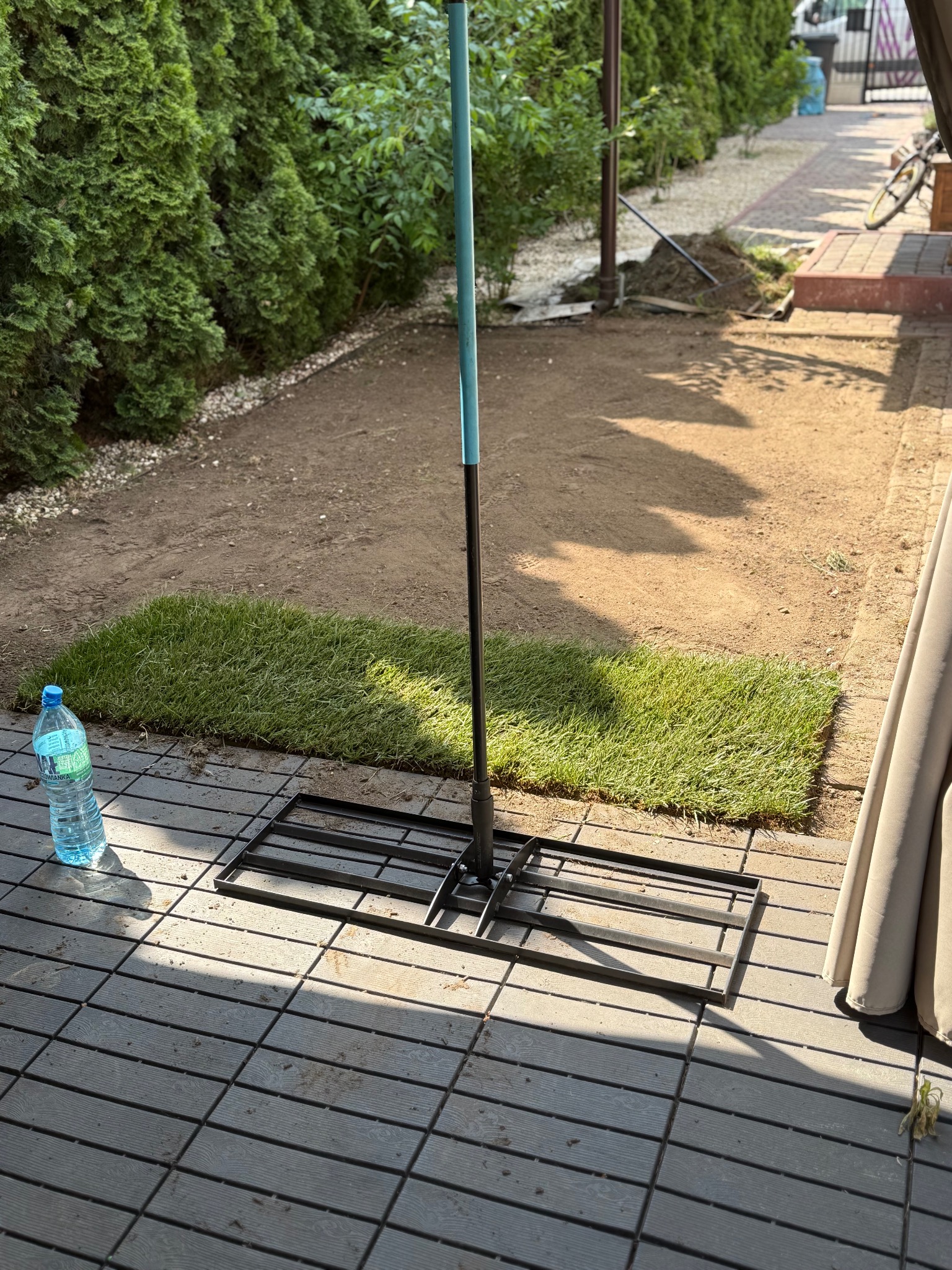 Fragment układania trawy z rolki w ogrodzie, widoczny fragment rolki trawy, narzędzie do jej układania oraz butelka wody. W tle zielone tuje.