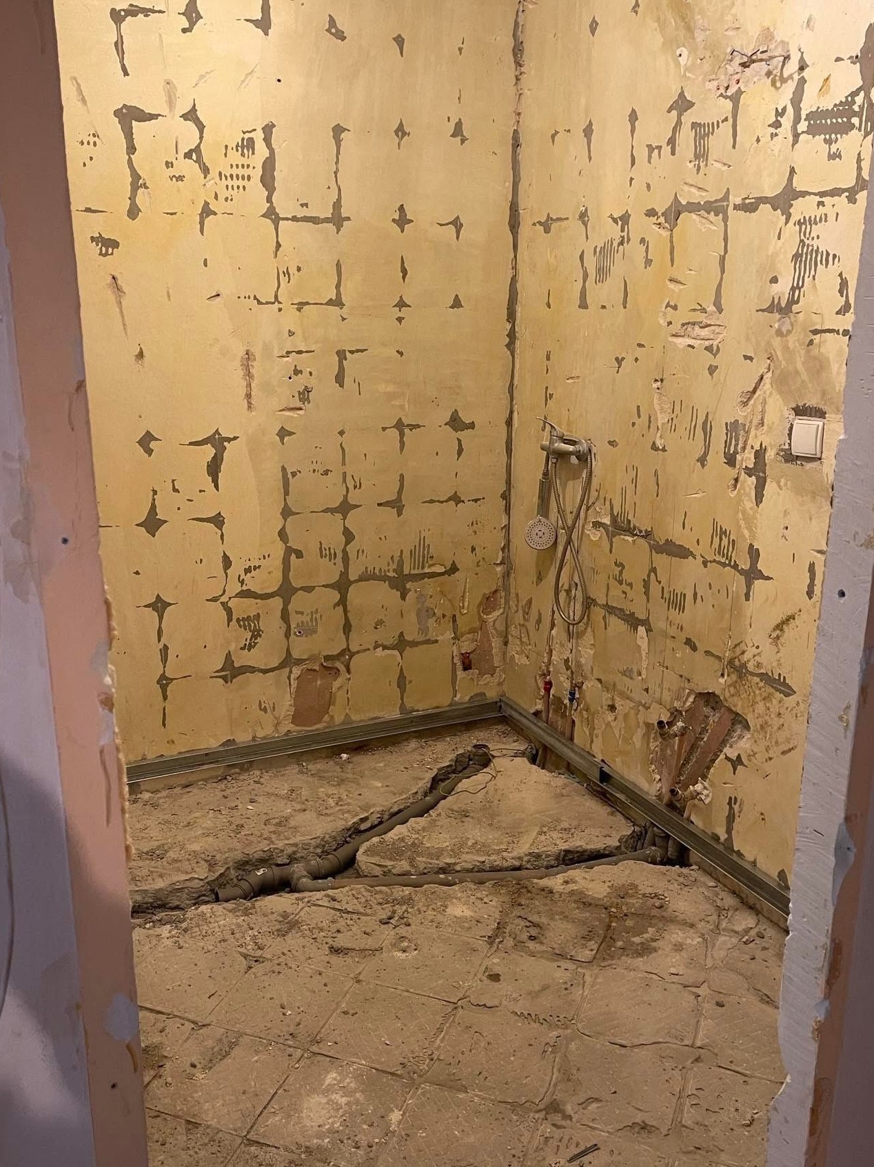 Wnętrze łazienki w trakcie remontu: zerwane płytki, odsłonięte rury, fragment armatury. Ściany z widocznymi śladami po demontażu, surowy stan przed wykończeniem.