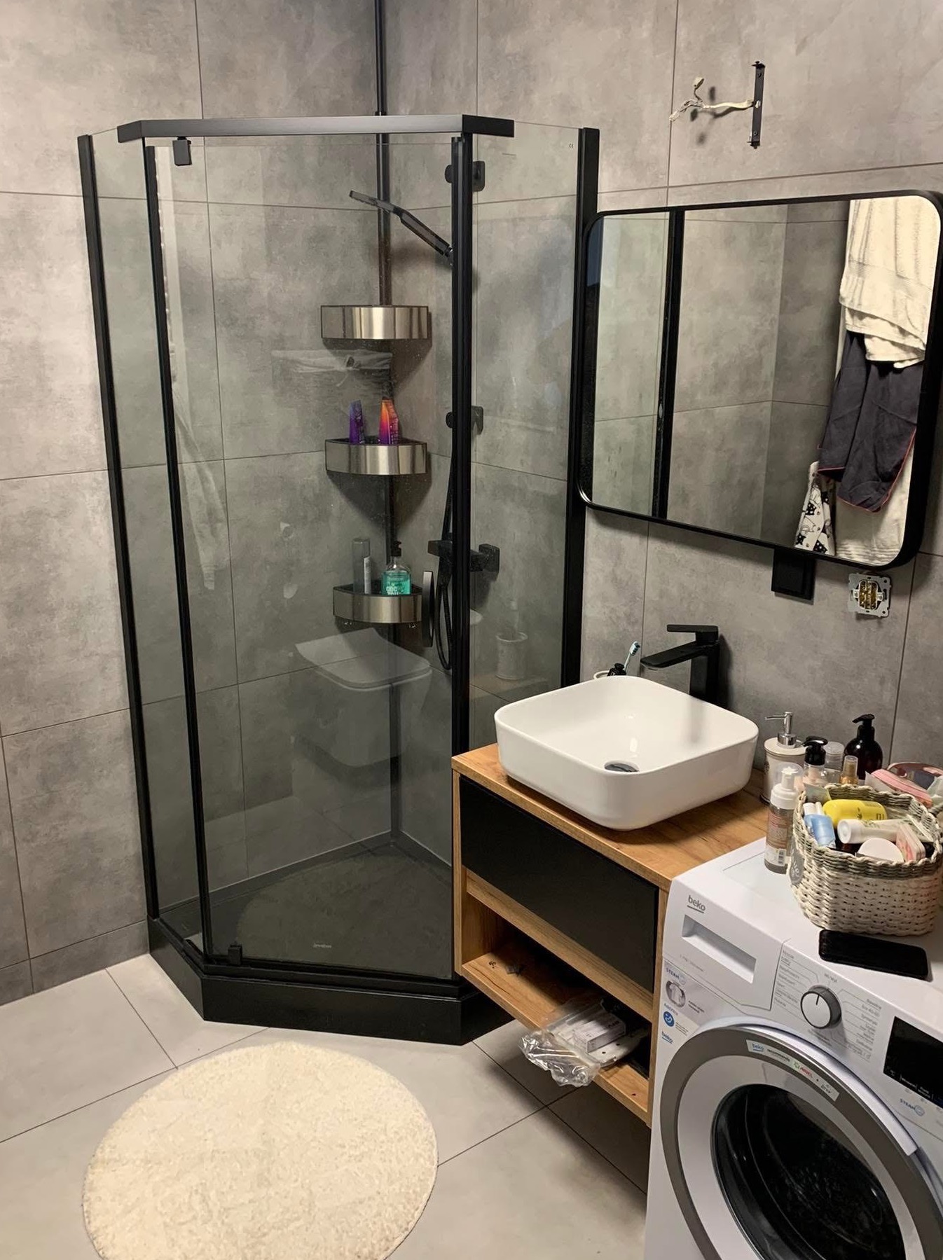 Nowoczesna łazienka z kabiną prysznicową w czarnej ramie, umywalką nablatową i pralką. Szare płytki i minimalistyczny design. Widoczny fragment lustra i akcesoria łazienkowe.