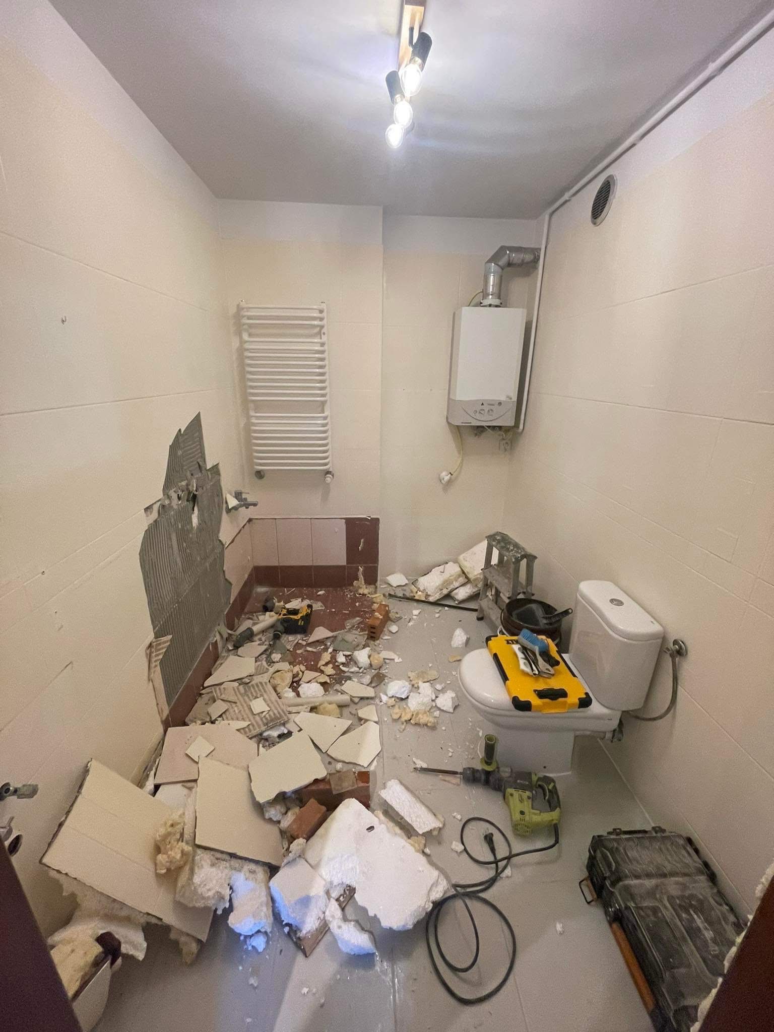 Demontaż płytek w łazience: uszkodzona ściana, gruz, narzędzia, toaleta. Widoczny bojler i grzejnik. Remont w trakcie.