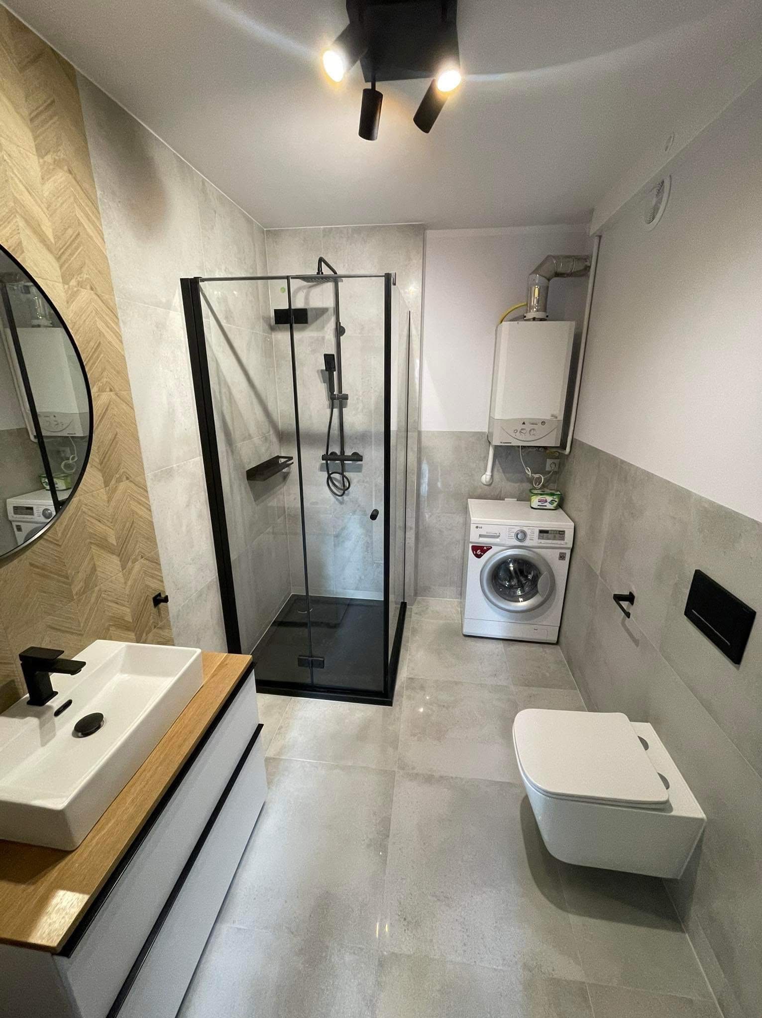 Nowoczesna łazienka po remoncie: kabina prysznicowa z czarną ramą, biała umywalka z drewnianym blatem, wisząca toaleta i pralka. Gres w odcieniach szarości.