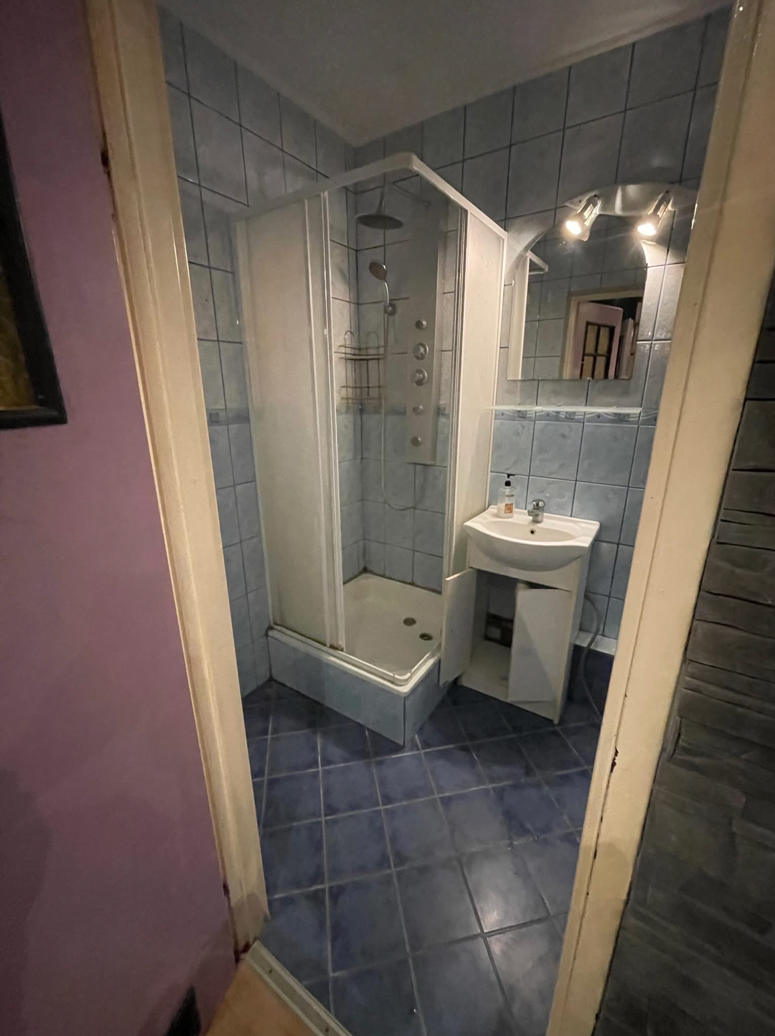 Niebieska łazienka z kabiną prysznicową, umywalką i lustrem. Widok zza otwartych drzwi.