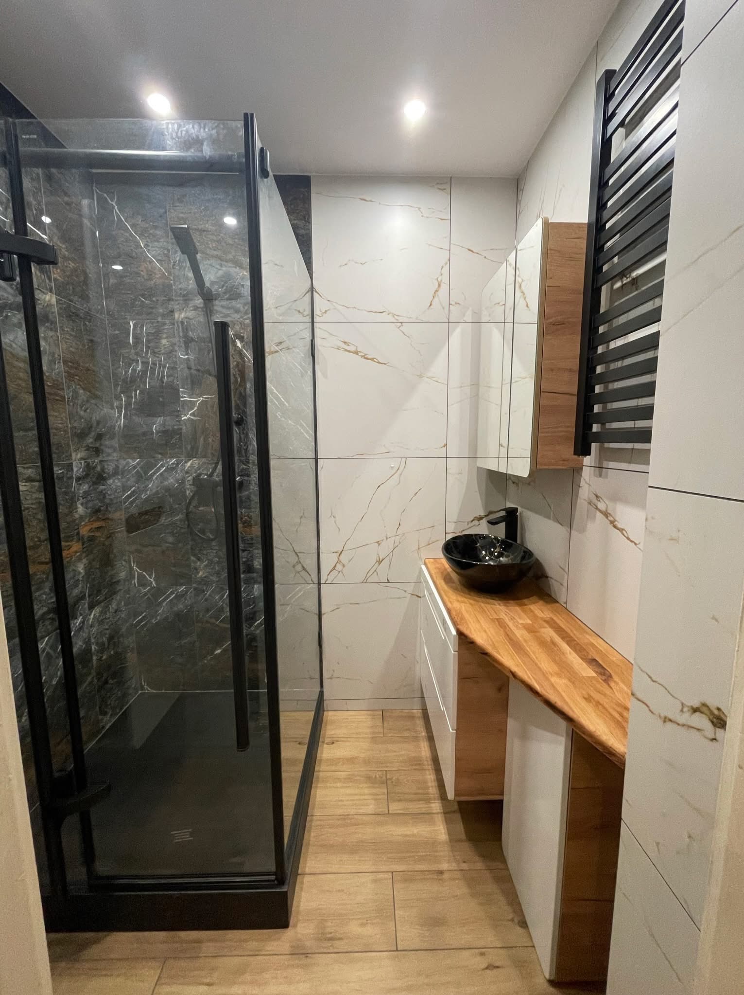 Nowoczesna łazienka z kabiną prysznicową typu walk-in, umywalką na drewnianym blacie i grzejnikiem drabinkowym w minimalistycznym stylu, wykończona płytkami imitującymi marmur.