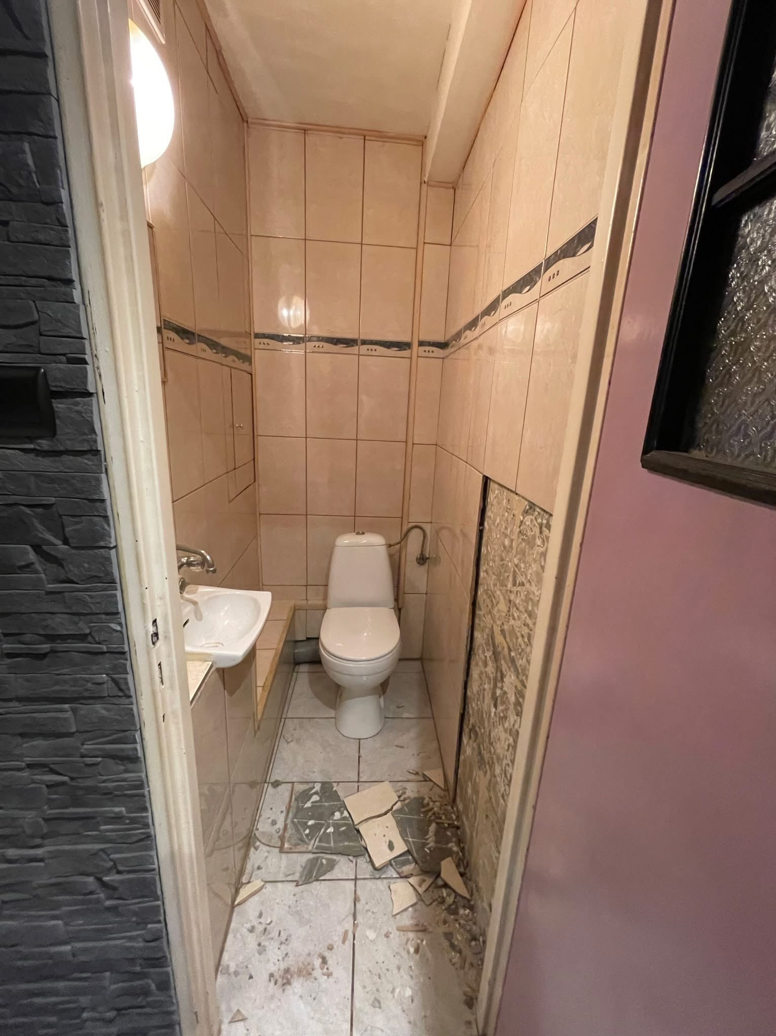 Łazienka w trakcie remontu. Widoczne uszkodzone płytki podłogowe i ściany, umywalka i toaleta. Ujęcie z perspektywy wejścia, oświetlenie górne.