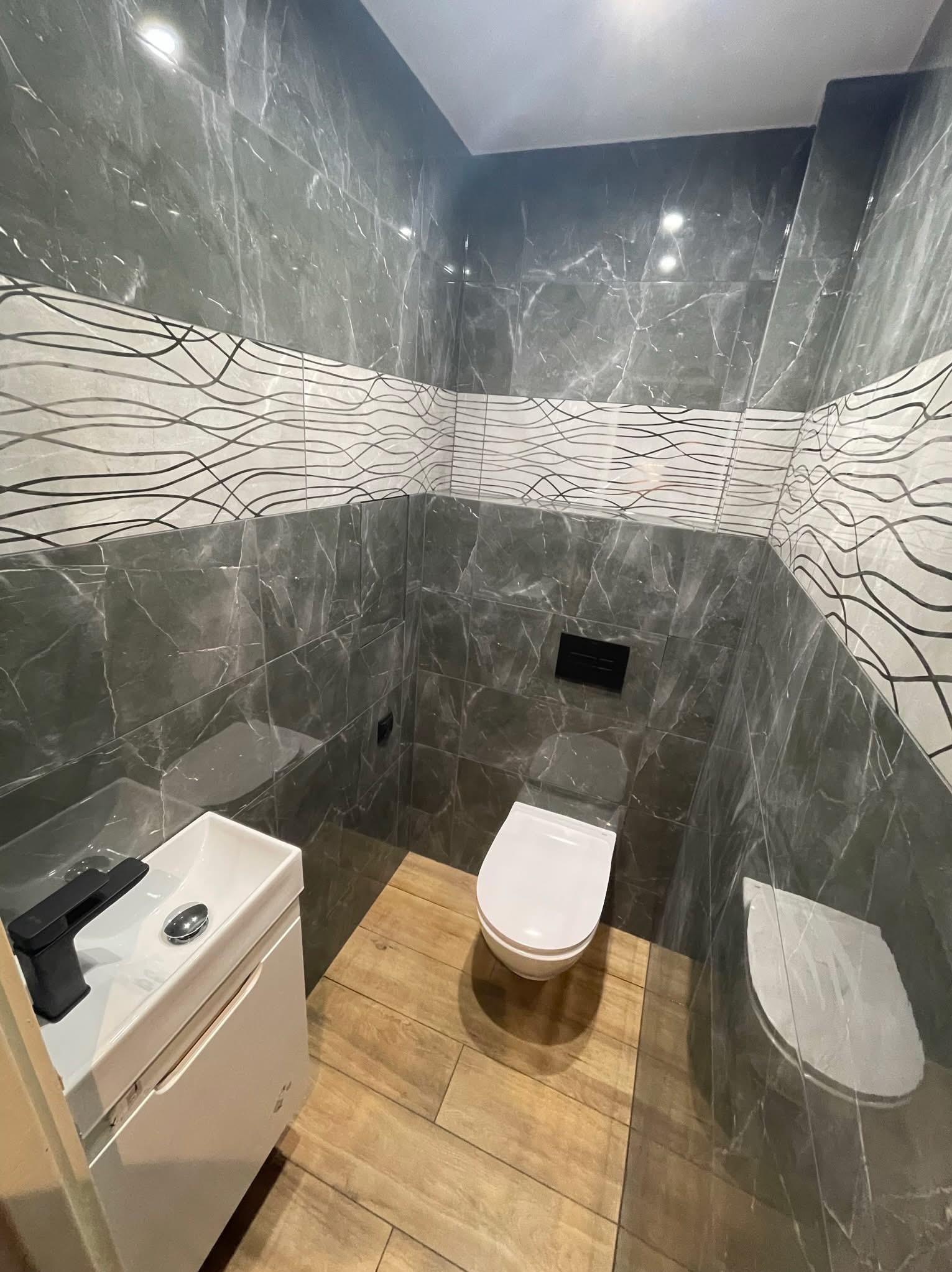 Nowoczesna toaleta z marmurowymi płytkami i drewnopodobną podłogą. Umywalka z czarną armaturą. Górny pas płytek z motywem fal. Całość w chłodnej tonacji.