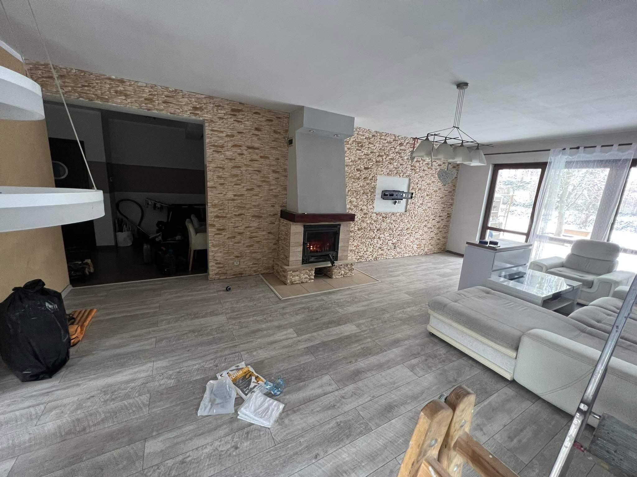 Salon po remoncie: kamienne ściany, kominek z paleniskiem, szare panele podłogowe i meble w jasnych barwach. Widoczny fragment drabiny malarskiej.
