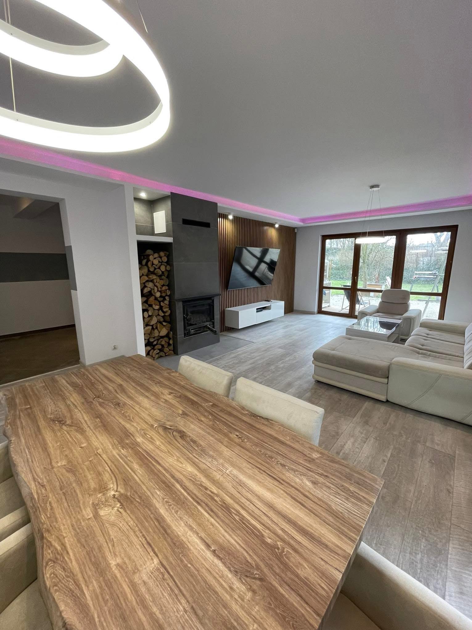 Nowoczesny salon z drewnianym stołem o nieregularnym kształcie blatu, kominkiem z zapasem drewna i oświetleniem LED w kolorze różowym.